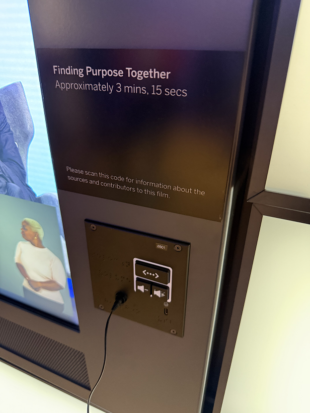 A wall panel beside a video screen reads "Finding Purpose Together", "Approximately 3 mins, 15 secs", above a UXP-A with a cable plugged into its headphone jack.