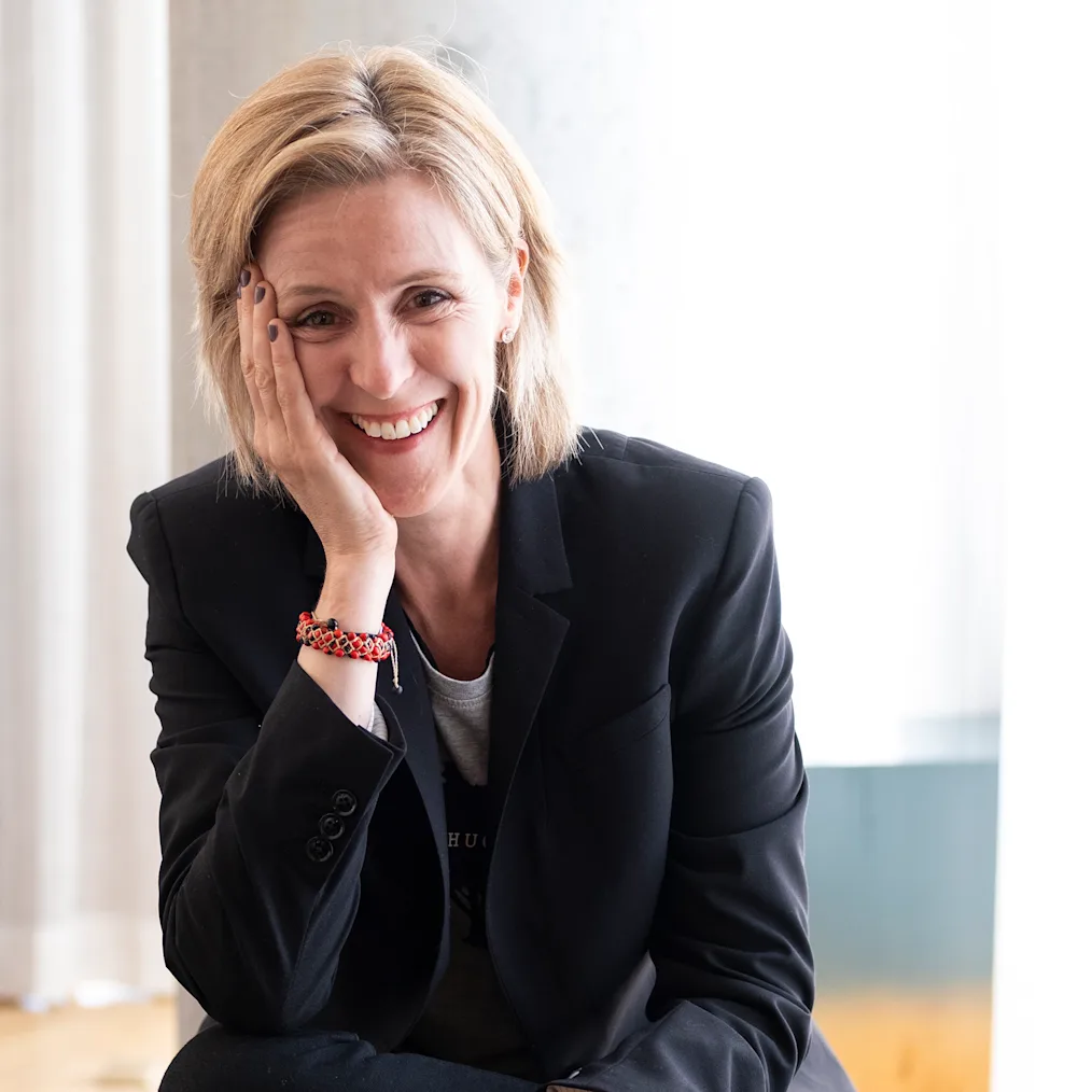 Ciara Byrne, an Irish woman with a light skin tone and blonde hair, smiles as she rests her hand on her face. She is wearing a black suit. 