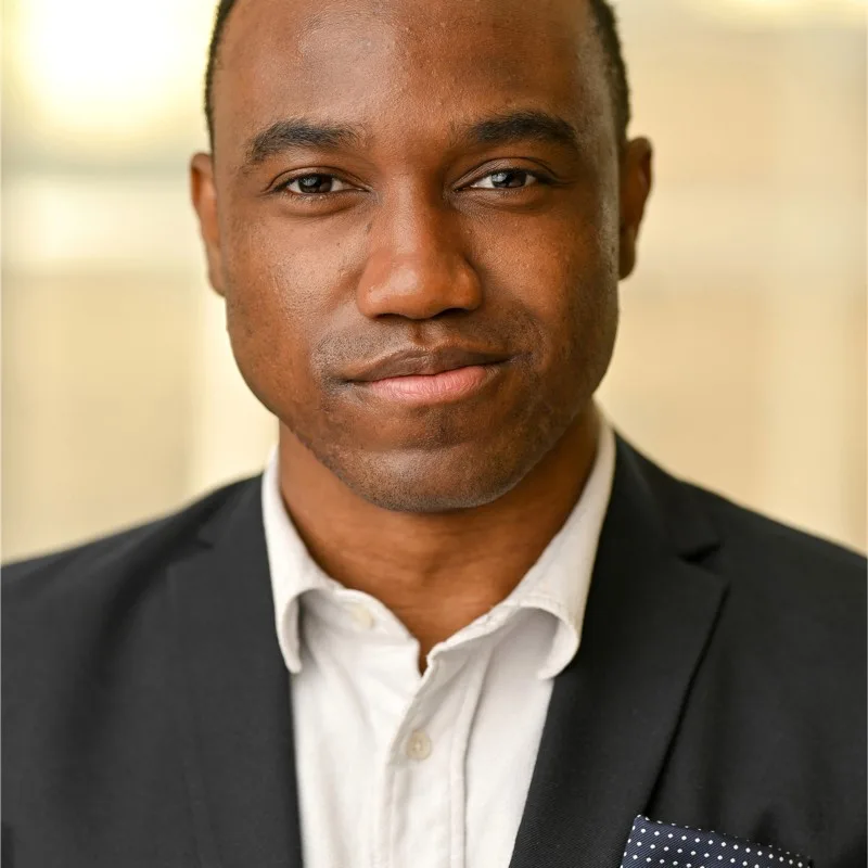 Zenge Simakoloyi, a Black man with a dark skin tone, holds a closed lip smile. He is wearing a black blazer and white shirt.