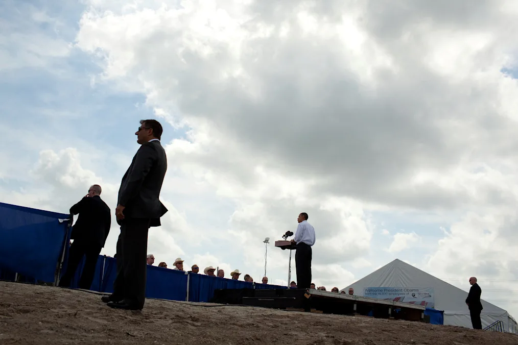 A belowimage of President Obama standing on dry dirt at a podium in front of a crowd of various people while men with light skin tones surrounds him in all black. 