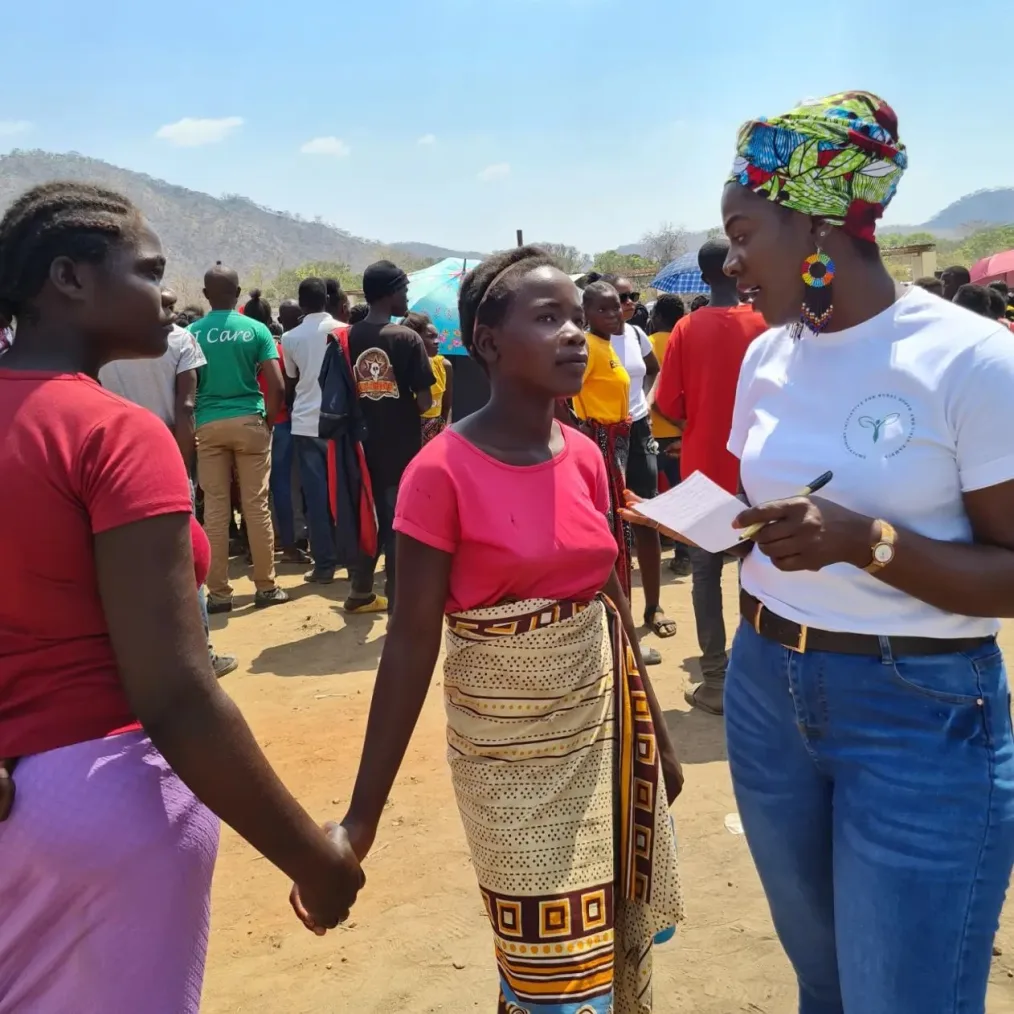 On a sunny day, Ireen Chikatula is pictured speaking with two adolescent girls in her community. The girls are holding hands. They have a deep skin tone and are wearing red and pink shirts. At least 20 people gather in the background.
