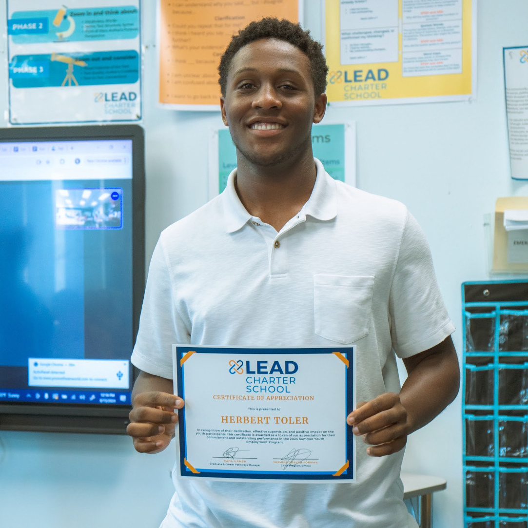 Herbert Toler, a young man with a dark skin tone smiles at the camera. He is holding a Lead Charter School certificate. 