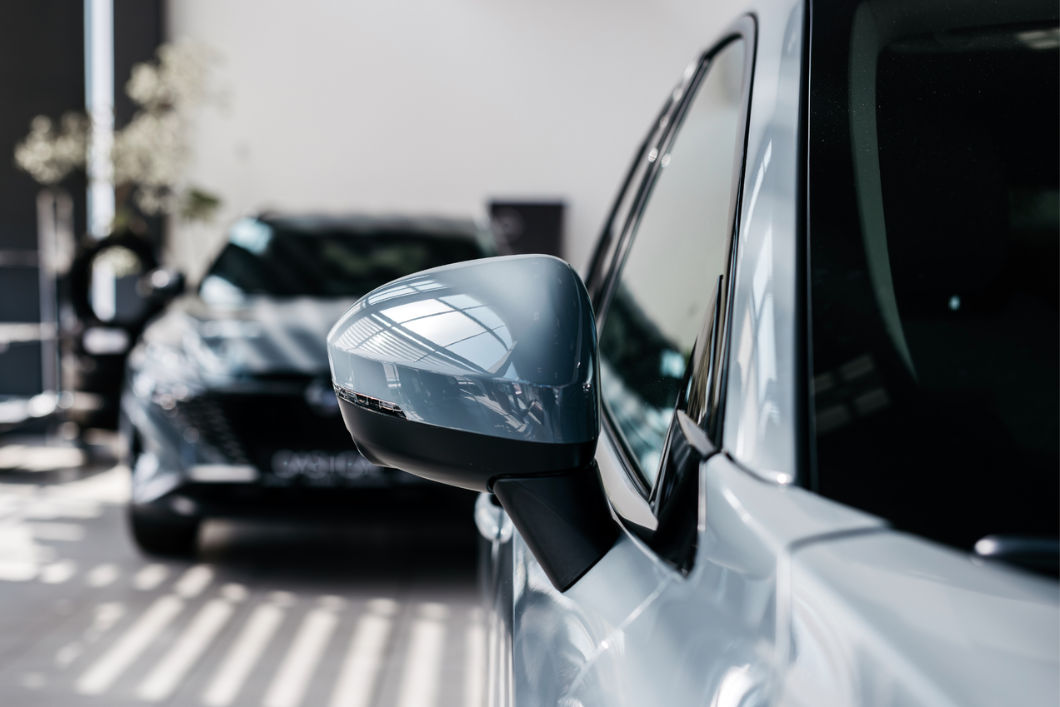 Närbild på sidospegel av silverfärgad bil i bilhall, med mörk bil i bakgrunden.