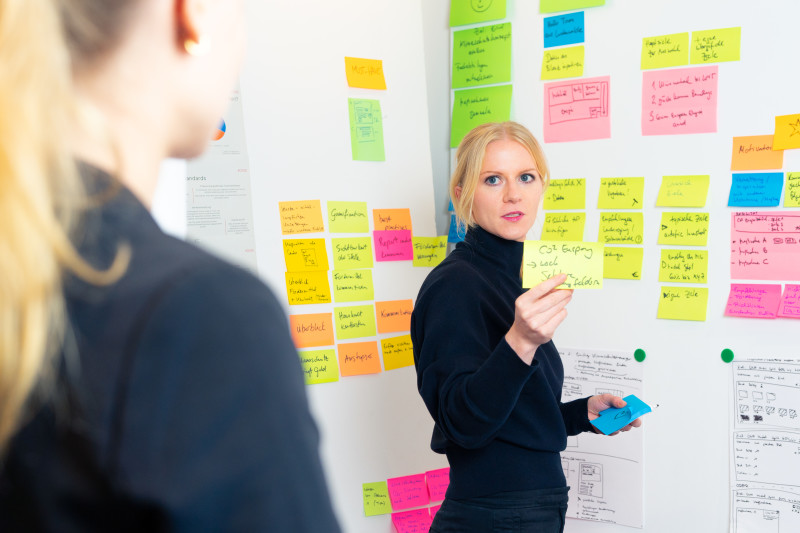 A woman pointing out a post-it during a workshop. Behind her, an entire wall of post-its can be seen.