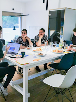 A busy working session with pretzels, croissants and drinks. Four people sit at a table in a bright office space, engrossed in conversation.