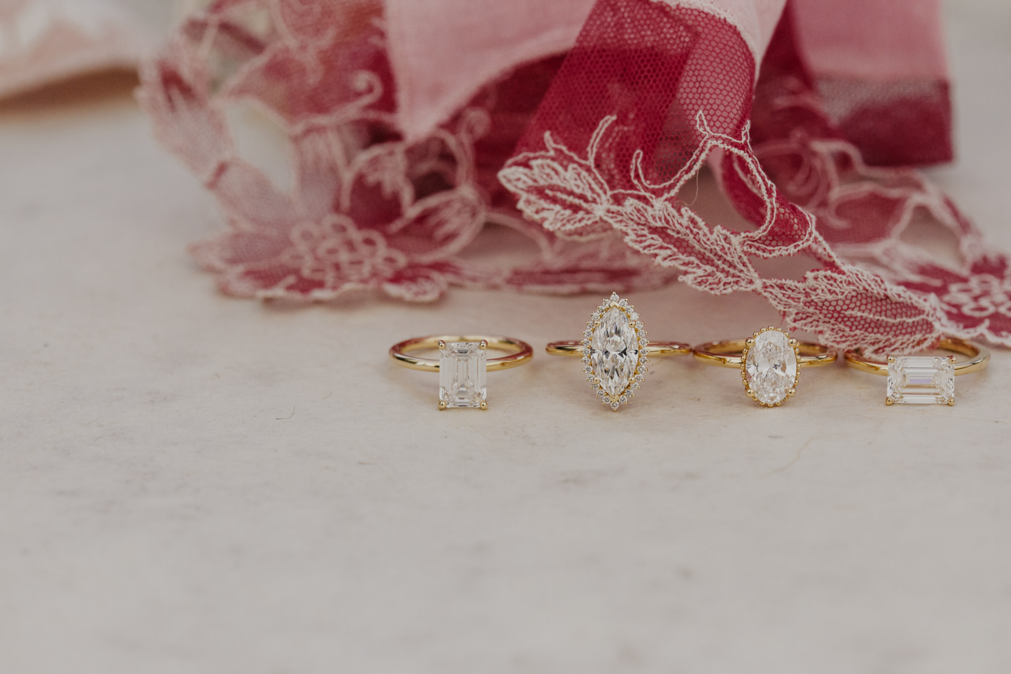 Four white diamond engagements rings in different shapes lined up with a pink ribbon background.