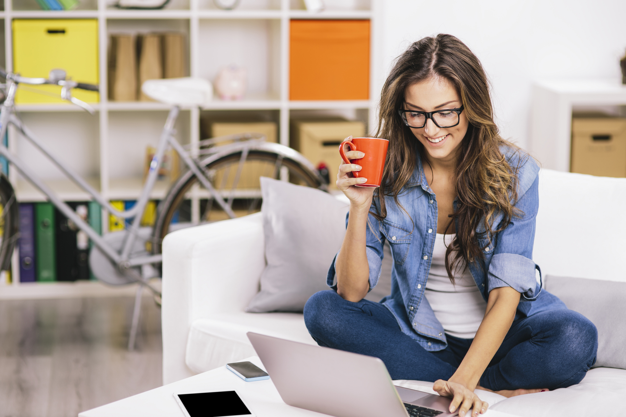 A smiling woman sits at home with a mug and a laptop.