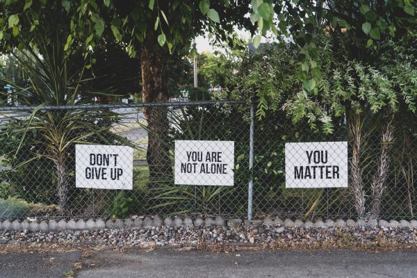 Three white signs behind a chain link fence in front of various trees. The signs say from left to right: "Don't Give Up", "You are not alone", and "You matter".