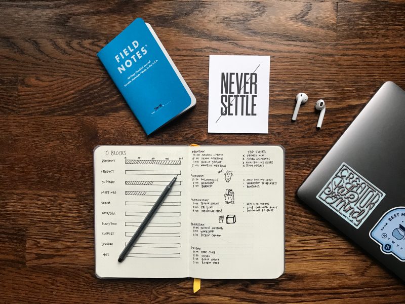 A dark wood desk background with a open journal on the desk open with sketches, above it is a light blue fields notes journal and to the right a white image that says Never Settle. To the right of the Never Settle is AirPods and a laptop in the corner. 