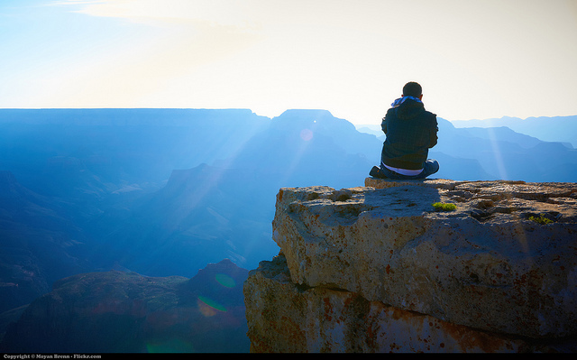A person sitting on a cliff facing the sun and a view of a mountain