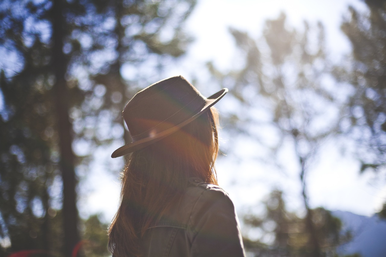 Woman with fedora hat on facing away from camera in front of trees.