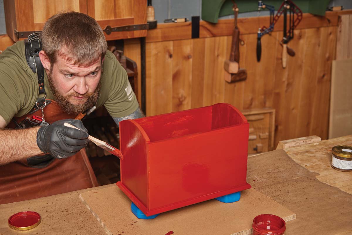 wood box covered in linseed oil red paint, man using paint brush to go over surface