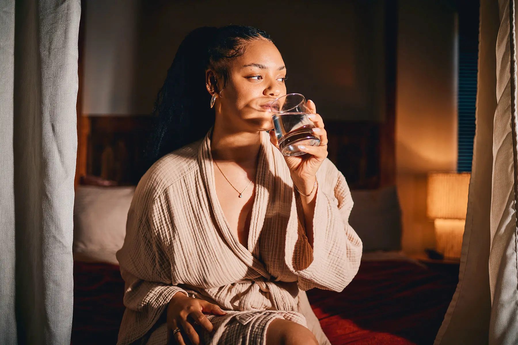 A Biracial woman dressed in a robe sitting on her bed, taking a pill with a glass of water. header