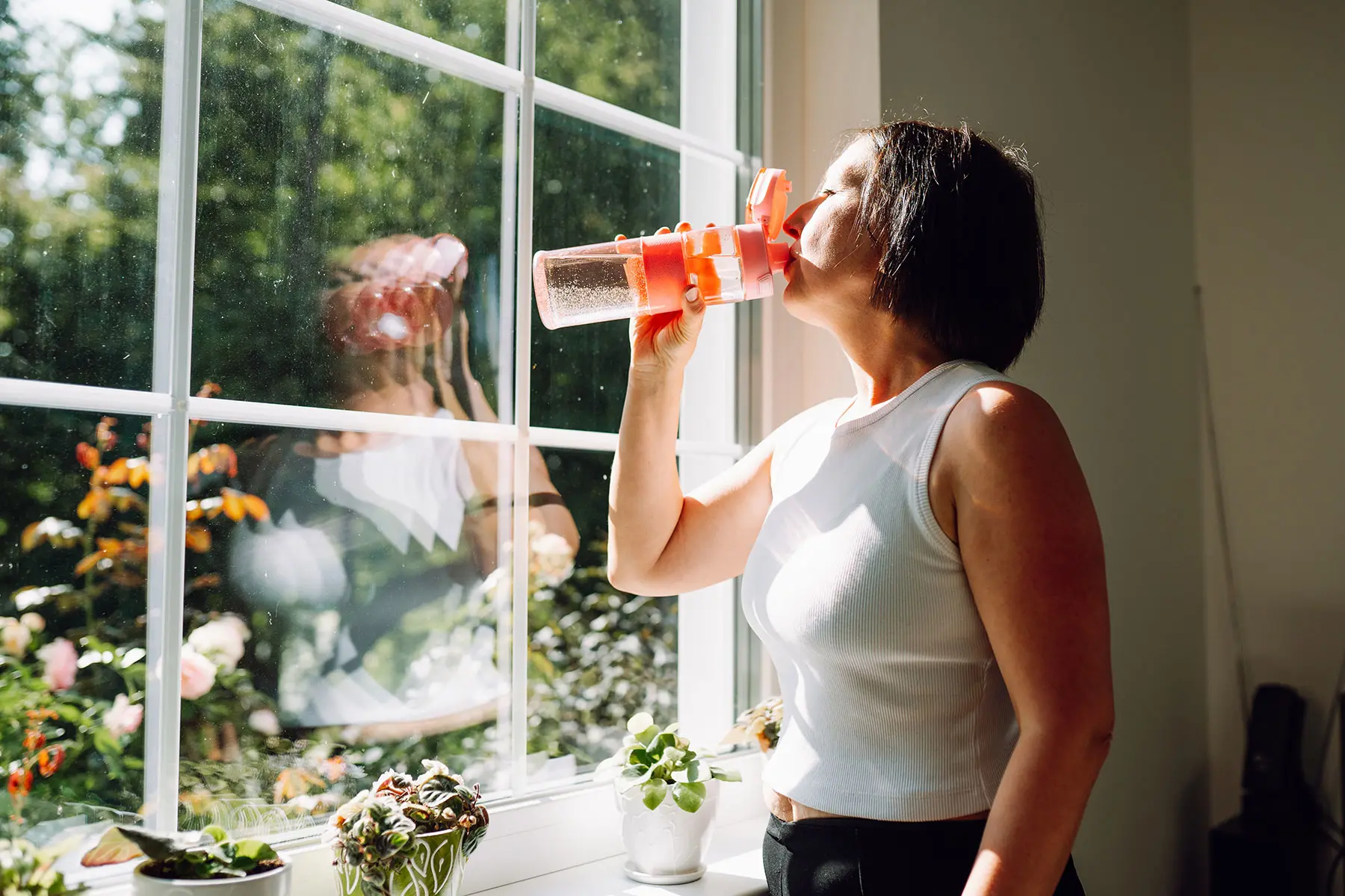 Woman standing in front of a large window in a sleeveless top, drinking from water bottle.