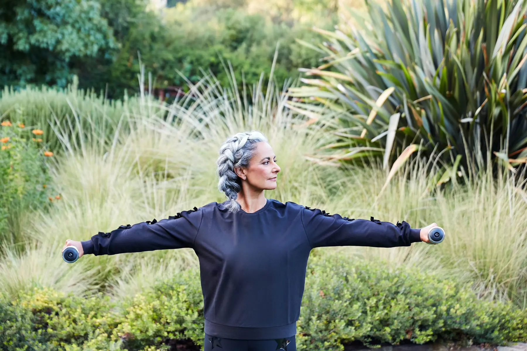 Fit woman with thick salt and pepper braid using weights to do arm exercises outdoors in lush landscape.