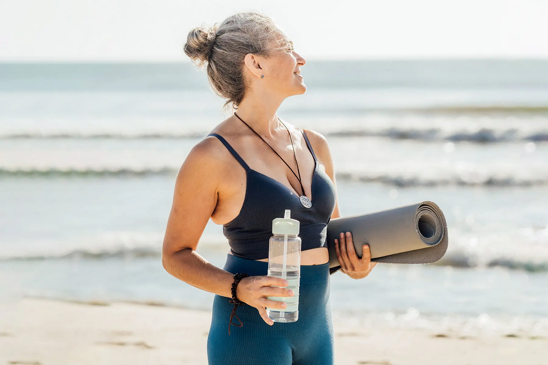 Woman standing at the beach wearing fitness clothing , looking away, holding a water bottle and yoga mat under her arm.