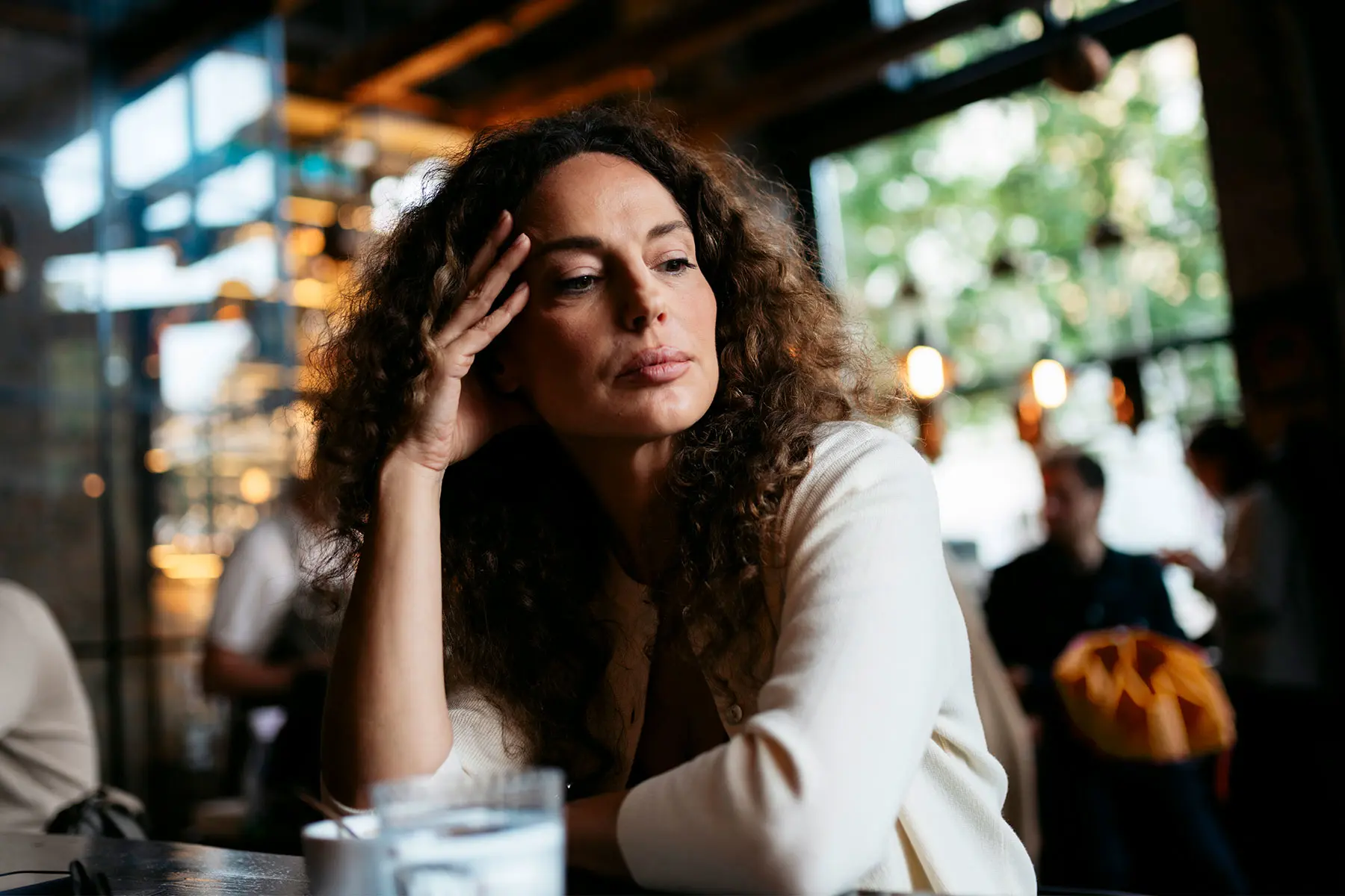 Mid adult woman with long curly hair, sitting at restaurant looking distracted, lost in her thoughts.