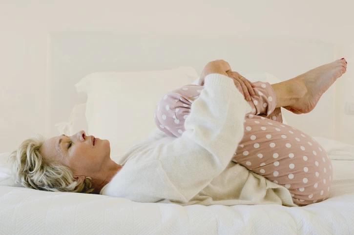 Mature blonde woman wearing polka dot pink pajamas in bed wrapping her arms around her legs, lying on her back. AW282