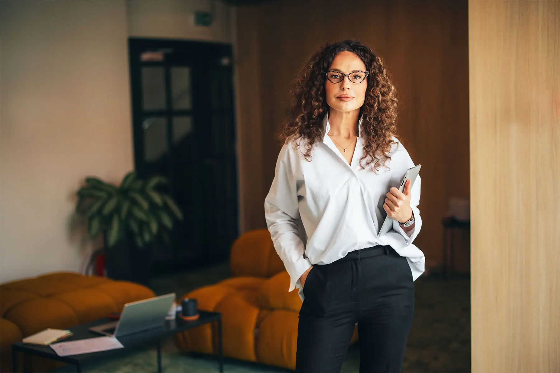 Confident middle-aged businesswoman in a white button down shirt, glasses and trousers looking at camera with a neutral expression, holding laptop in a warm toned office.header