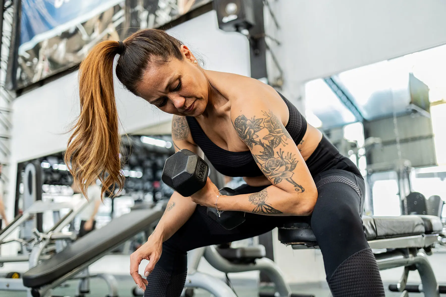 Mature Latin woman with tattoos is using free weights to exercise her biceps in a gym. header