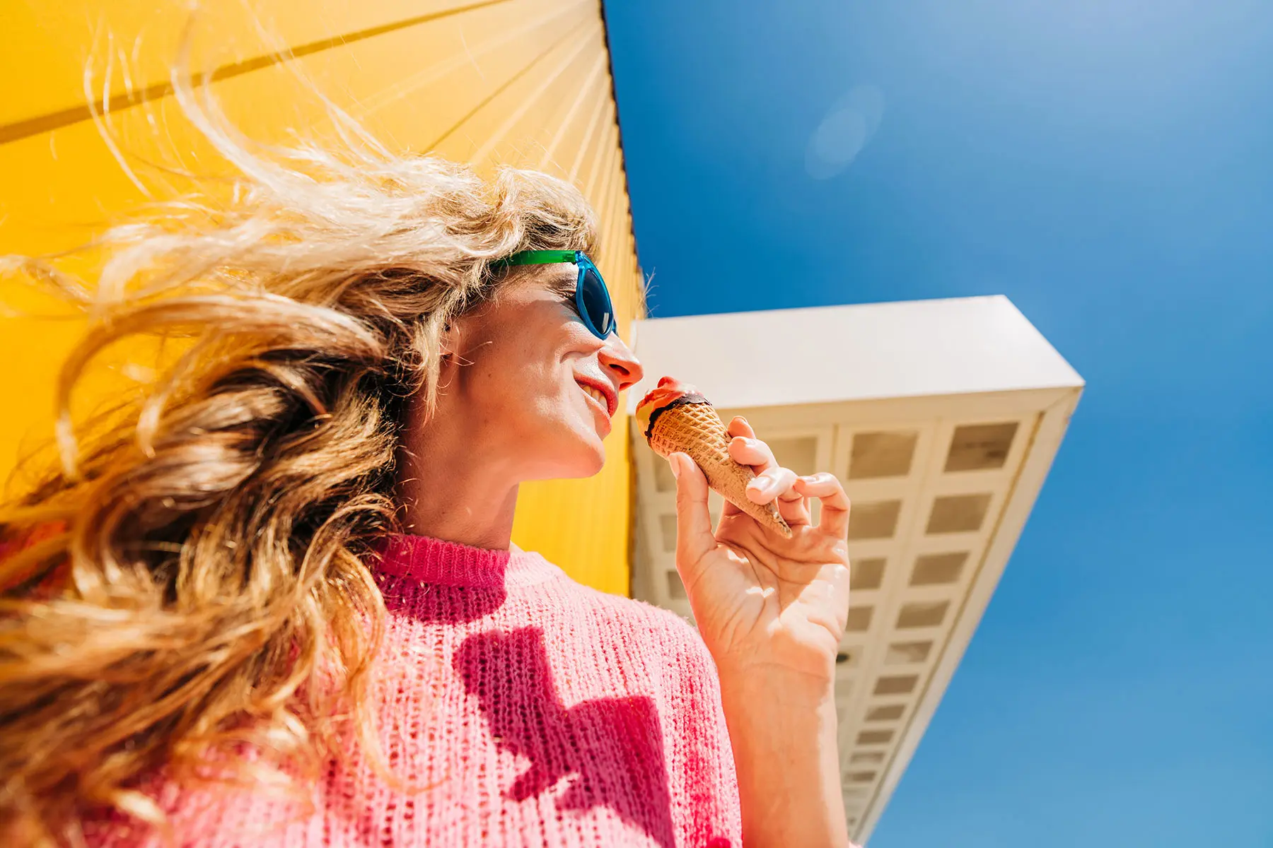 Low angle view of a woman with long blonde hair wearing a pink sweater and sunglasses eating an ice cream cone. header