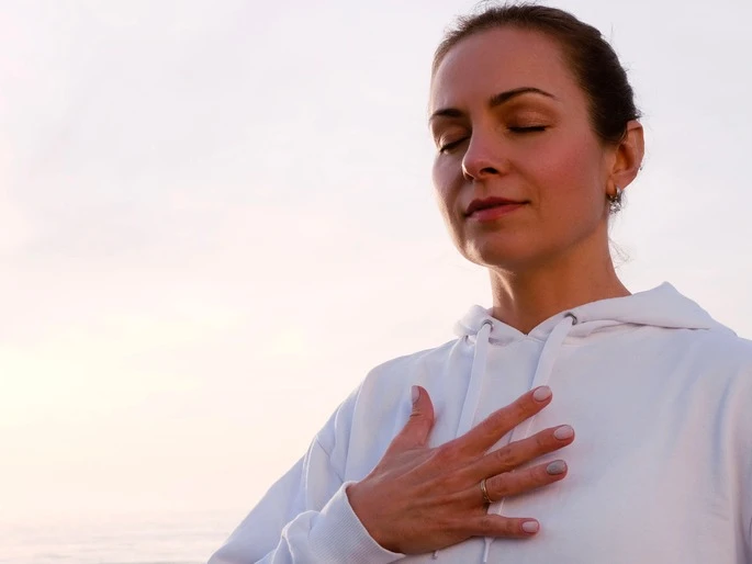Woman outdoors at sunset in hoodie with her hand over her chest, eyes closed. AW138