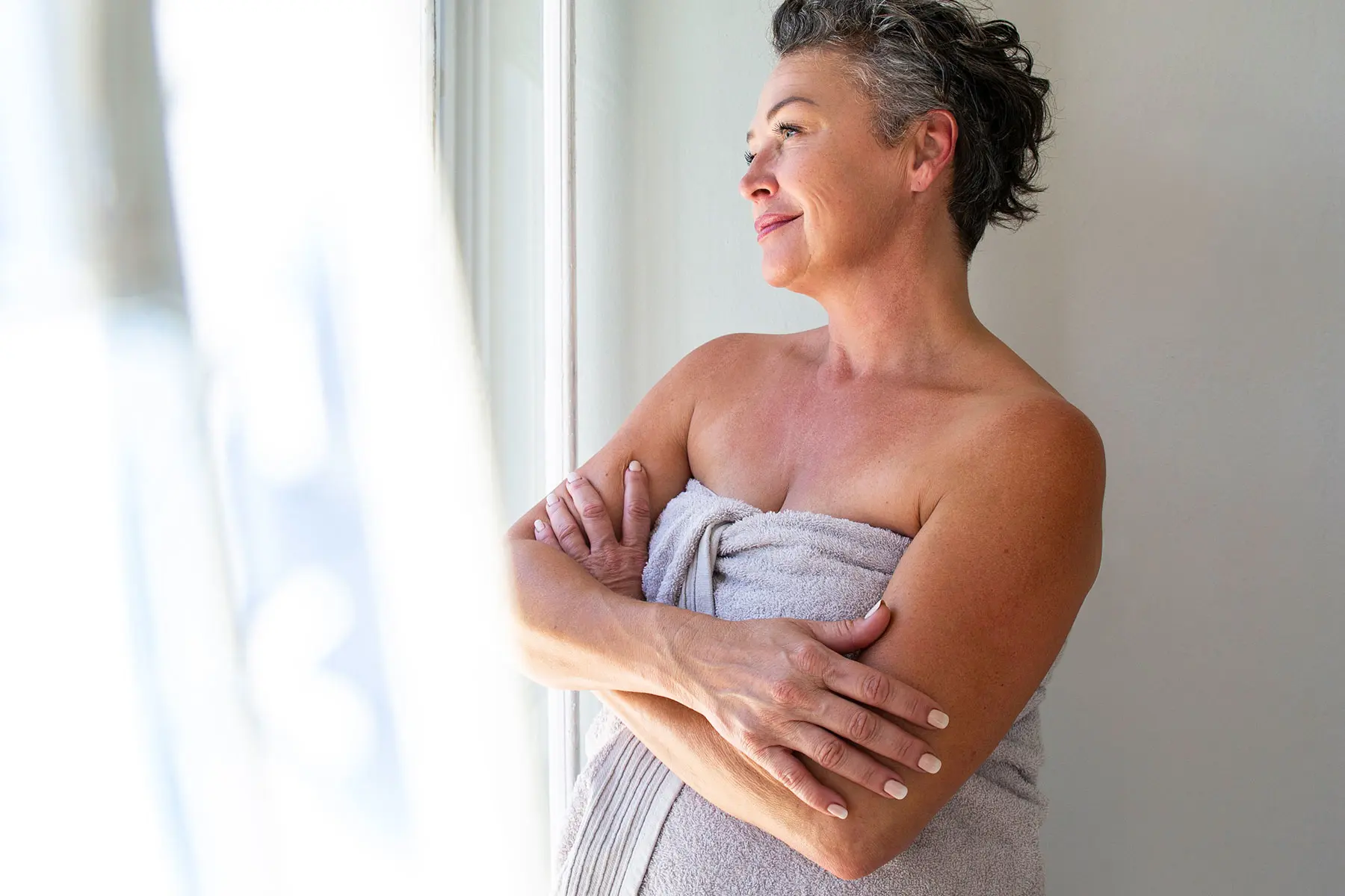 Middle aged woman standing in a bathroom, wrapped in a towel after using estradiol mist, and taking a shower. header