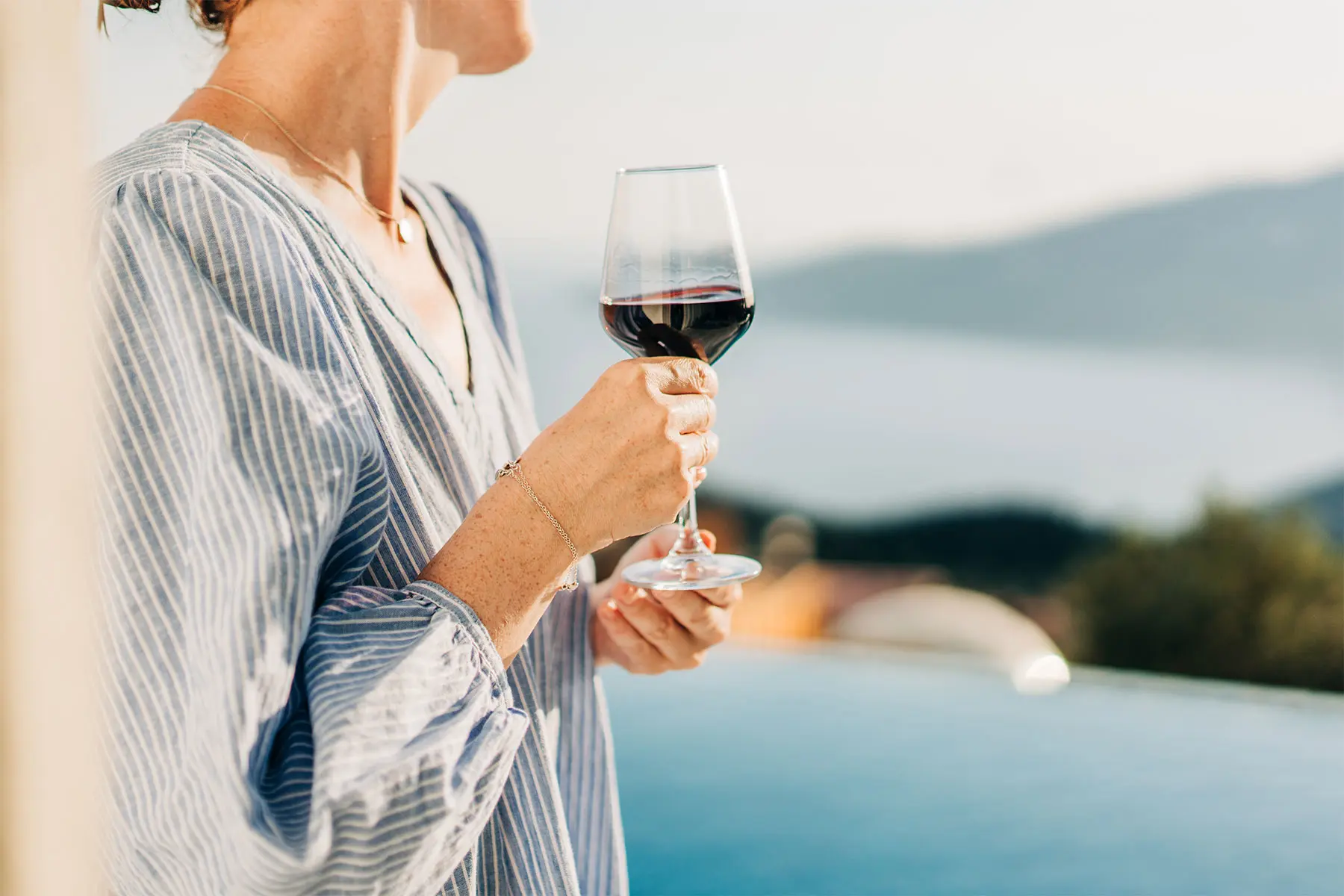 Torso crop of a woman in a striped blouse, outdoors holding a glass of red wine. top image