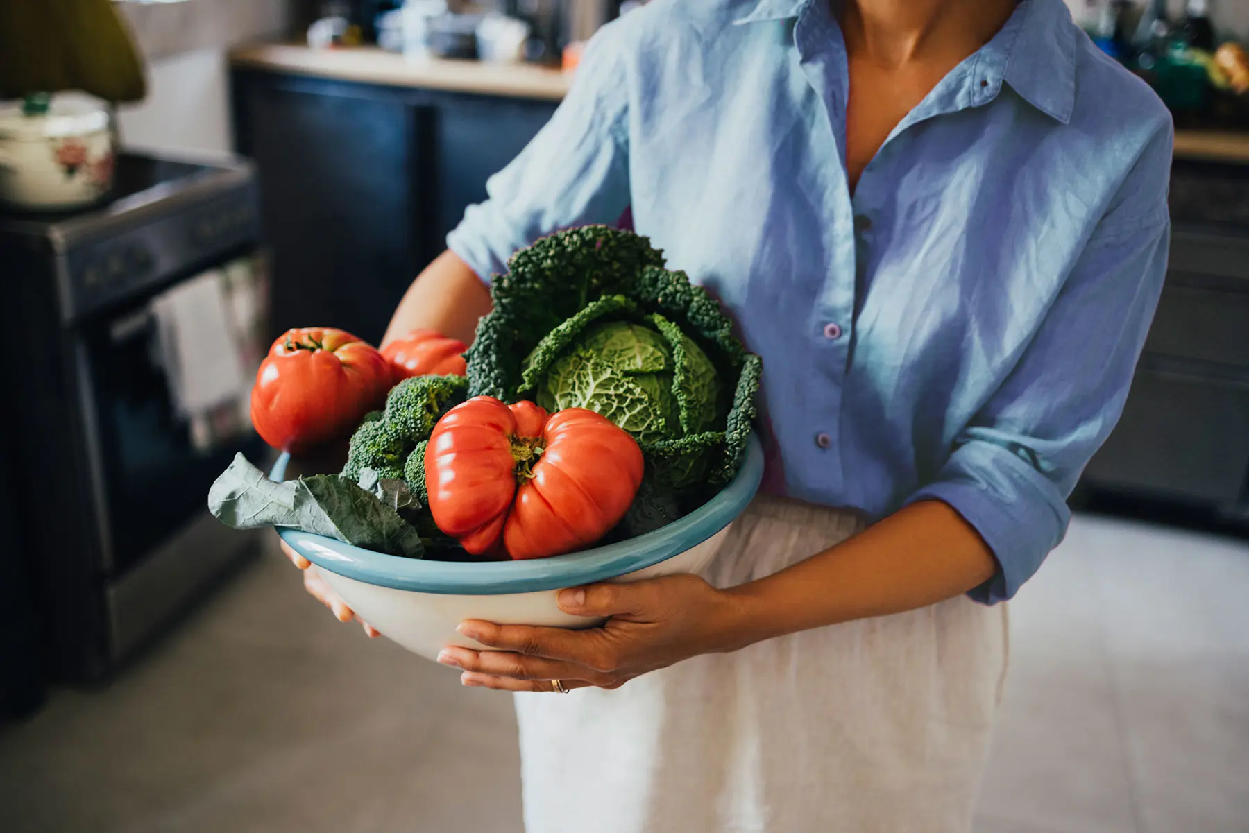 Cropped view of a woman in kitchen, wearing blue linen shirt carrying large bowl of fresh tomatoes, cabbage and broccoli (header)