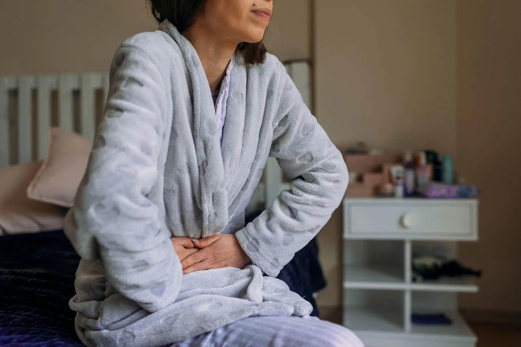 Middle-aged woman sits one the edge of her bed in pale blue bathrobe, with her hands gripping her abdomen in discomfort