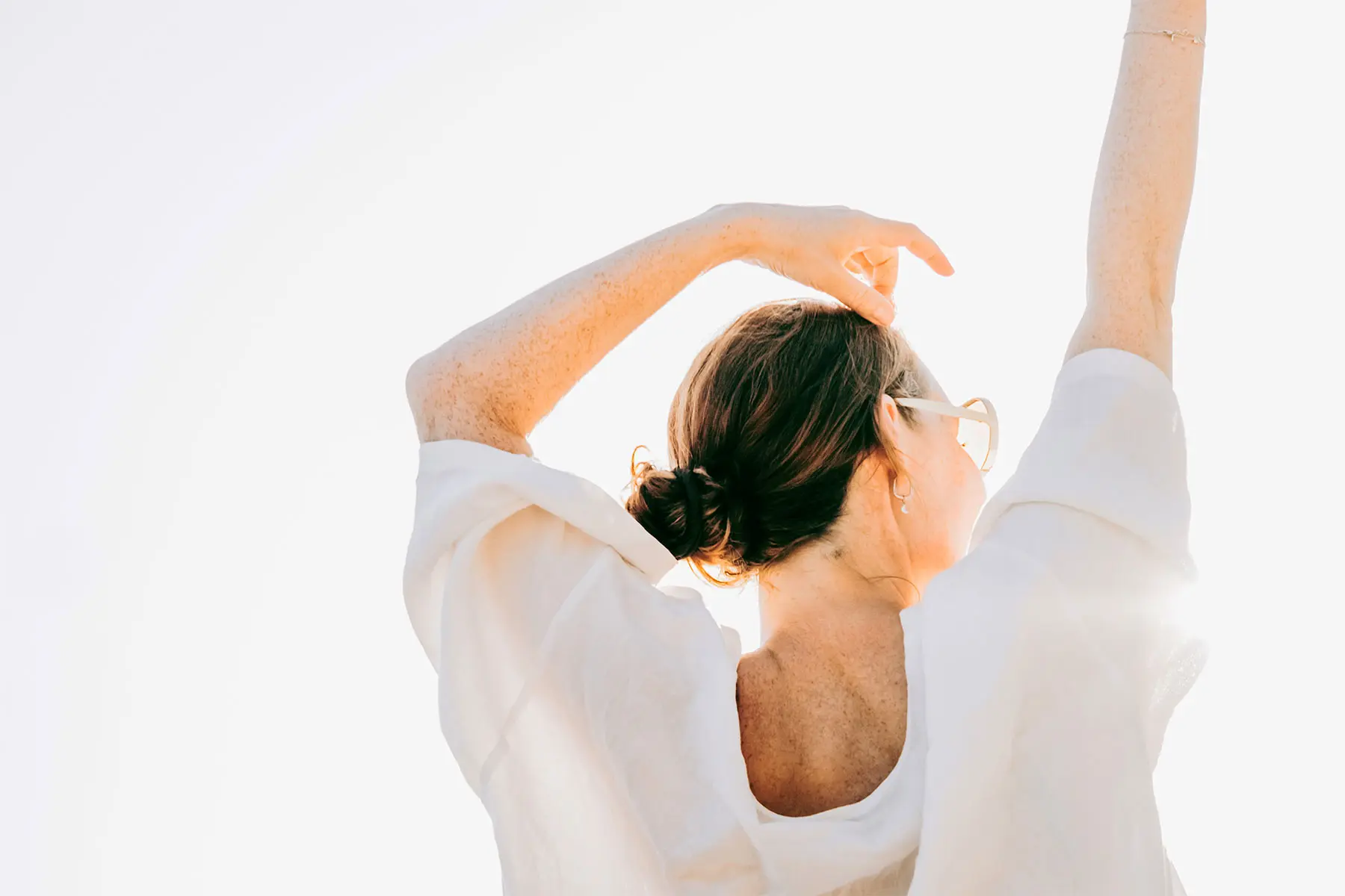 Woman in natural fiber blouse and sunglasses seen from behind, with her arms lifted above her head.