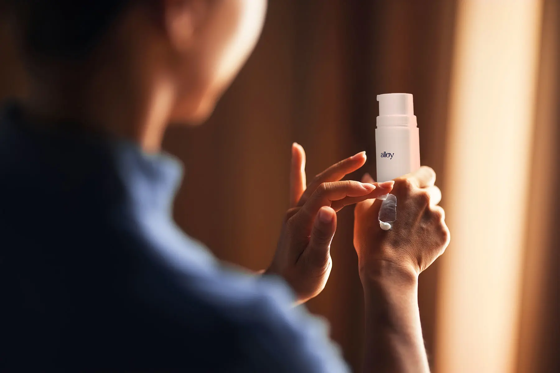Over the shoulder view of a woman holding Alloy M4 skin cream pump, applying cream to hand. header