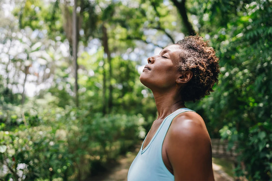 Mature woman standing on path in lush greenery wearing blue fitness tank top pausing to breath with eyes closed (inline)