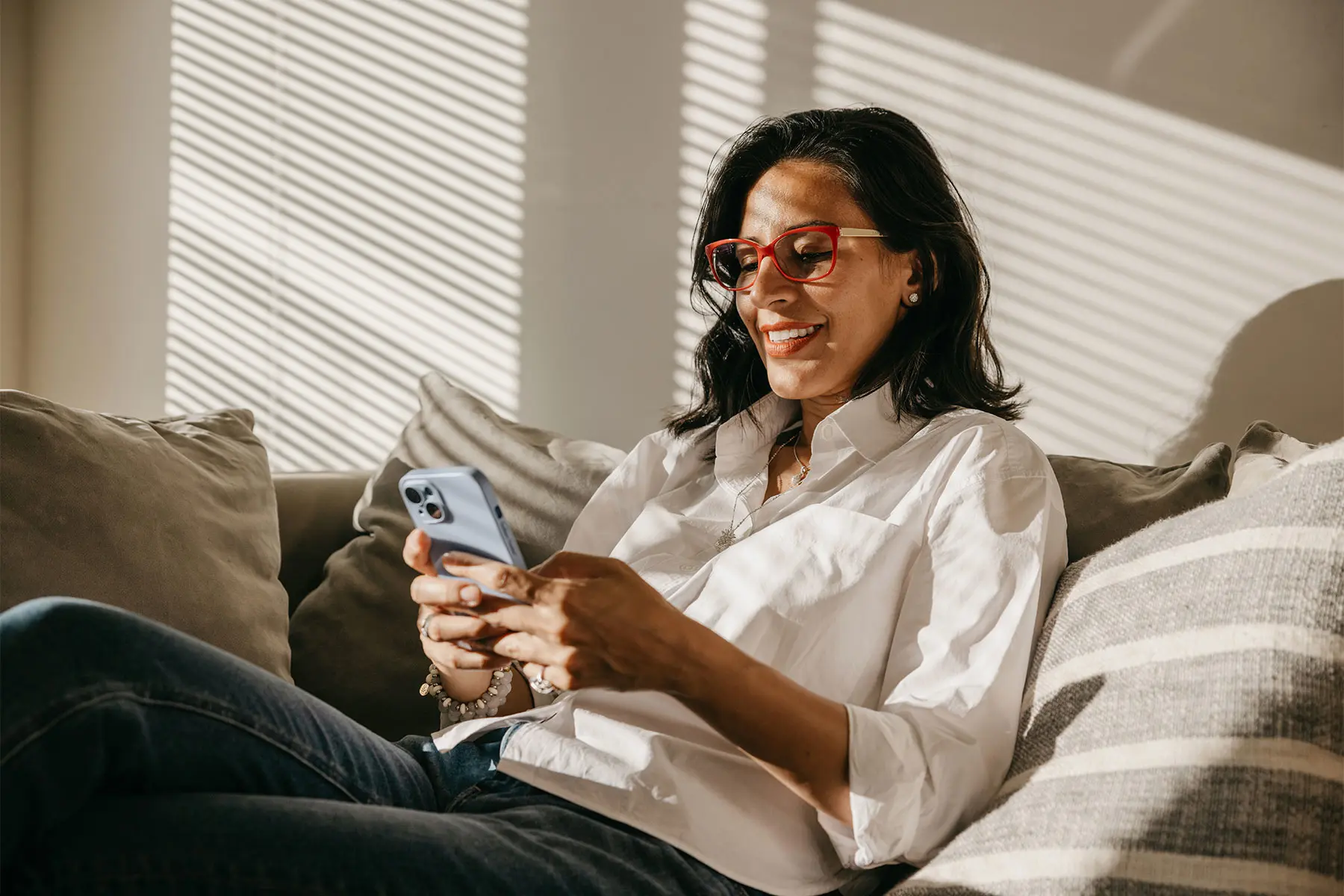 Middle aged woman wearing eyeglasses, sitting in the sunlight on a sofa, checking a hormone tracking app on her phone. header