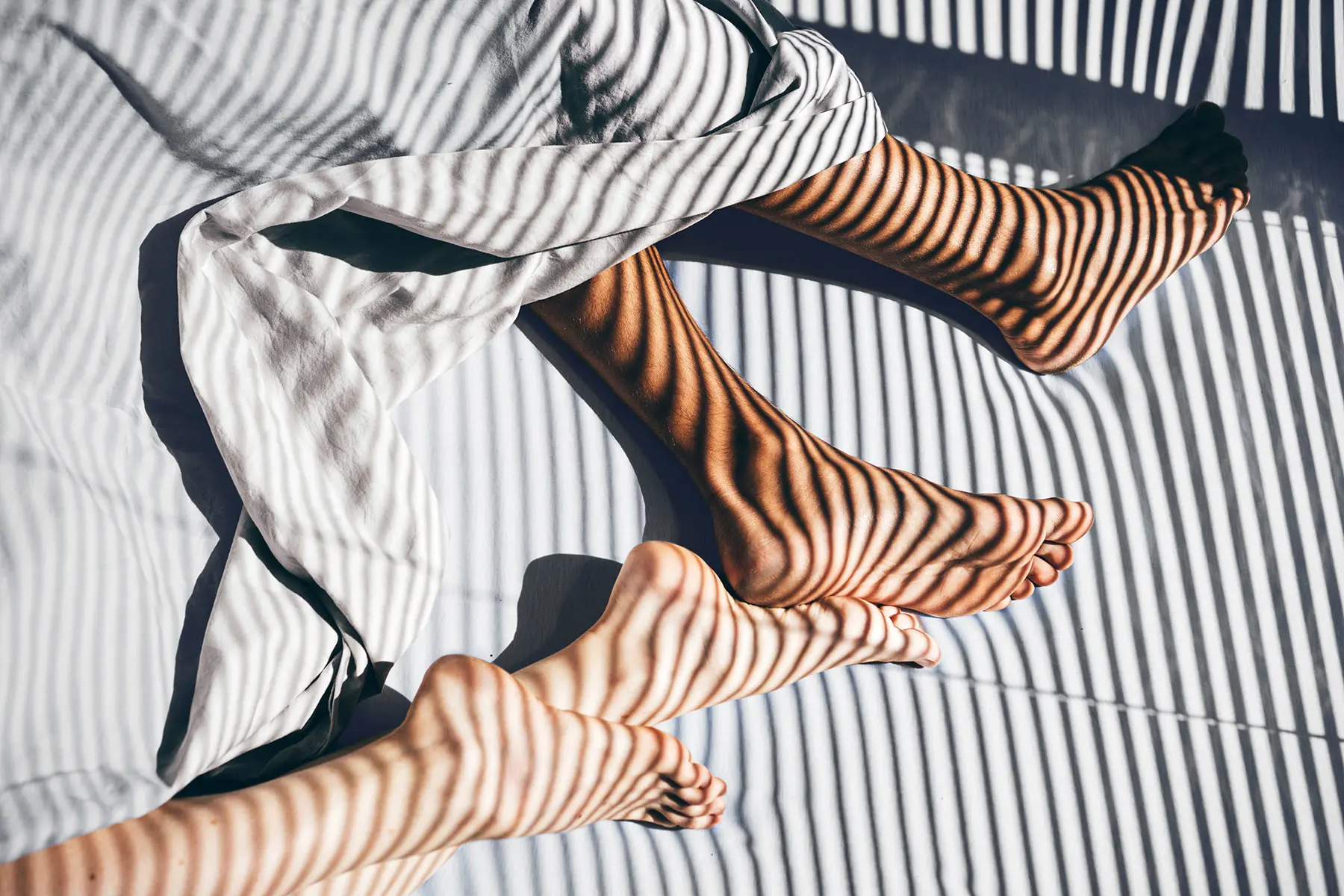 Unrecognizable couple 's legs intertwined in sheets from above cast in shadows from window blinds. header