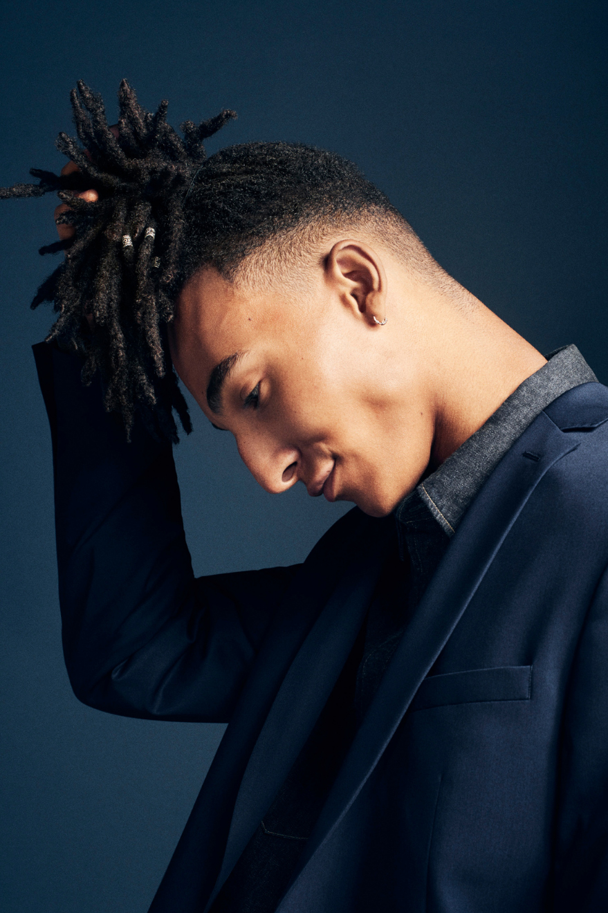 A young man with long dark hair pinned up holding his bun and looking down standing sideways to the camera.
