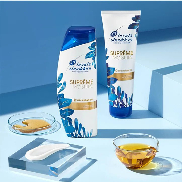 A Supreme Moisture Shampoo and Conditioner bottles standing on a blue surface next to the oil in the glass bowls and a creamy streak on the piece of glass.