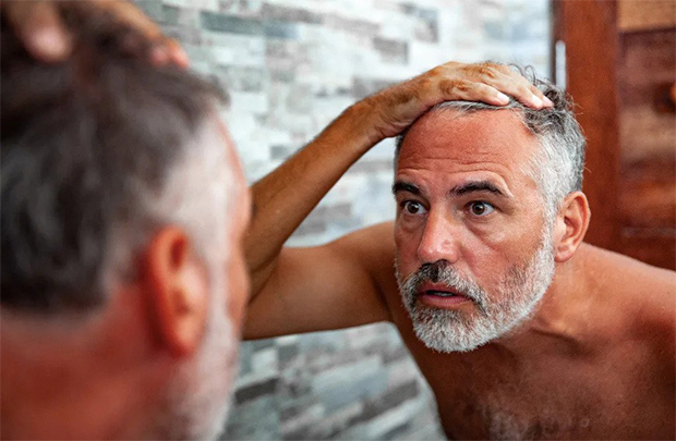 An old man with grey hair and beard looking at his hairline in the mirror.