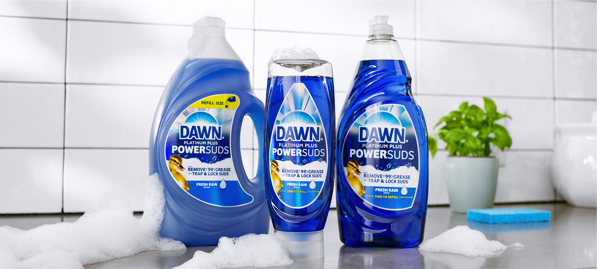 Productos PowerSuds sobre una superficie blanca con espuma a su alrededor, con azulejos blancos y una flor en el fondo.
