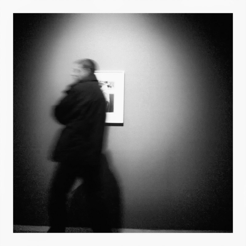 Black and White pinhole-style photo of person passing by a photograph in a gallery.