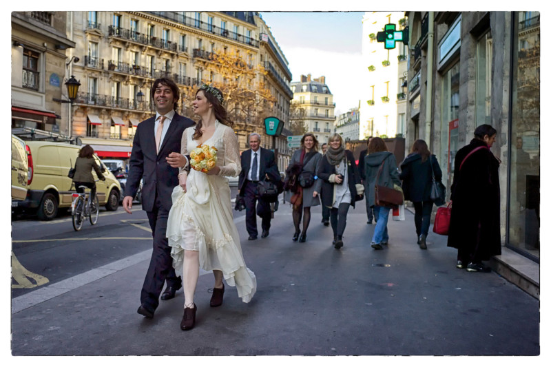Colour photo of newly married couple walking down Parisien street. The woman is carrying yellow bouquet of flowers.