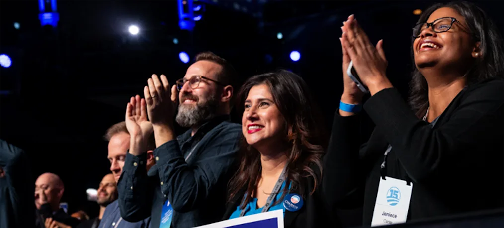Attendees applaud during an indoor event, standing behind a barrier with “Hello Chicago!” displayed in front.
