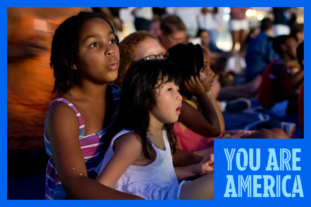 Children sit together outdoors at dusk, watching a performance with focused expressions as a crowd gathers behind them.