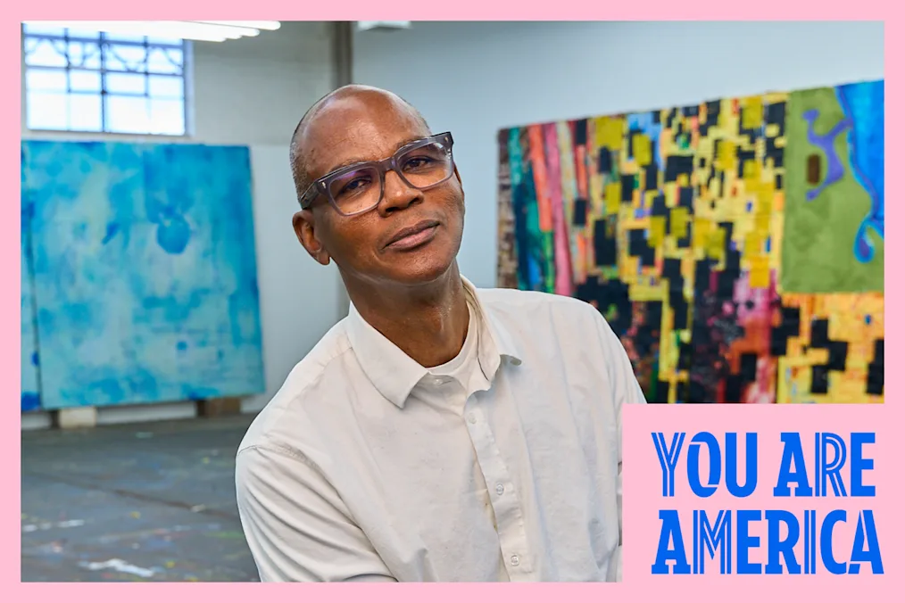 Artist Mark Bradford sits in his studio surrounded by large, colorful abstract paintings.