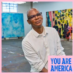 Artist Mark Bradford sits in his studio surrounded by large, colorful abstract paintings.