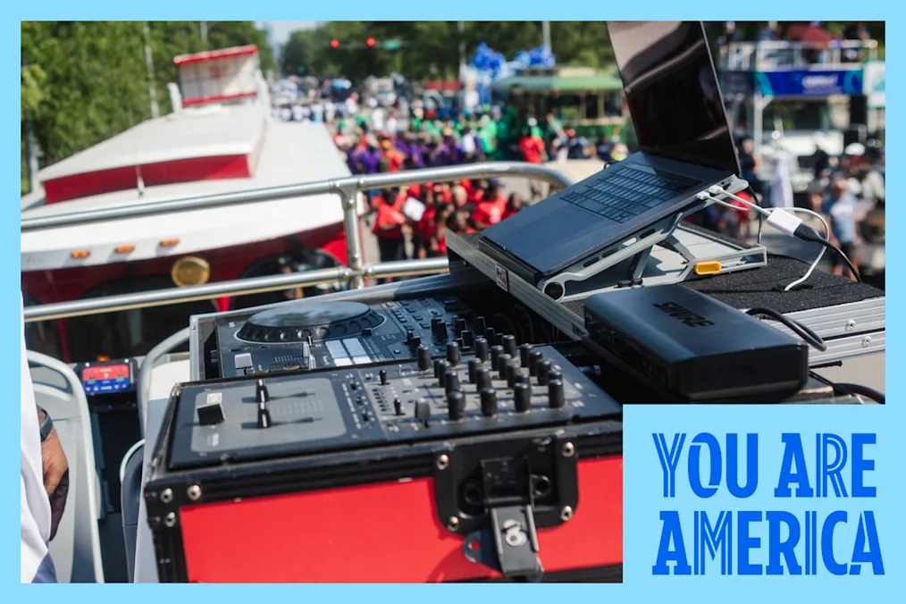 A DJ stands at a mixing booth on an open-top setup, pointing toward the crowd during a daytime street event.