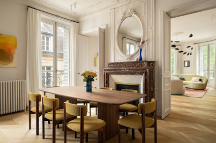 Dining room with walnut table, mustard upholstered chairs, ornate marble fireplace and oval mirror in a classic Parisian apartment.