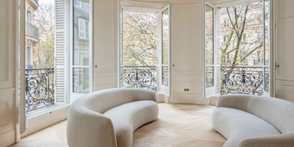 Sunlit Paris 7th arrondissement Haussmann sitting room with curved sofas, herringbone wood floors, and tall French doors opening to wrought-iron Juliet balconies with leafy street views.