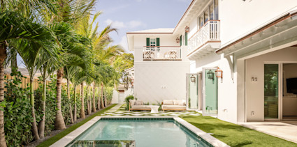 Backyard pool framed by palm trees and checkerboard pavers, with a white two-story home and breezy lounge seating.