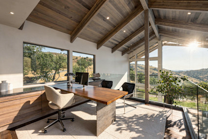 Sunlit home office with a wood desk, beamed ceiling, and floor-to-ceiling windows framing rolling oak-covered hills.